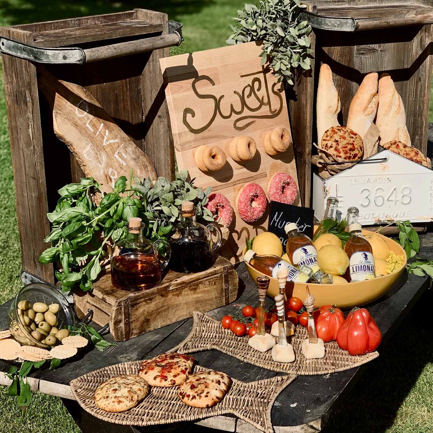 Rustikaler Tisch mit Holzkisten, Brot, Oliven, Kräutern und Limonade im Freien bei Sonnenschein
