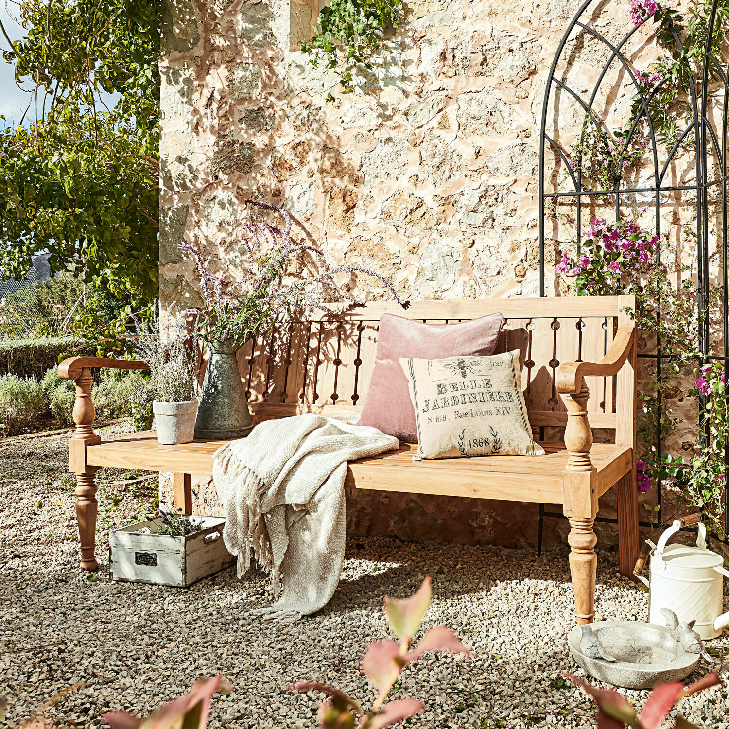 Bank Loudoun Helle Holzbank im Garten mit Kissen und Decke auf Kiesboden vor Steinmauer und Pflanzen