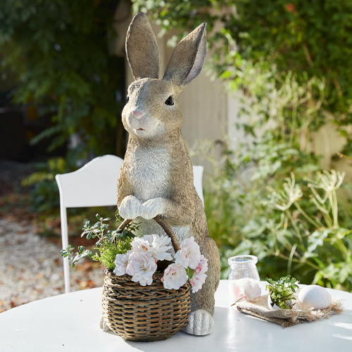 Dekorativer Hase aus Kunststoff in Naturfarben mit Weidenkorb und Blumen auf dem Gartentisch