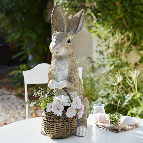 Dekorativer Hase aus Kunststoff in Naturfarben mit Weidenkorb und Blumen auf dem Gartentisch