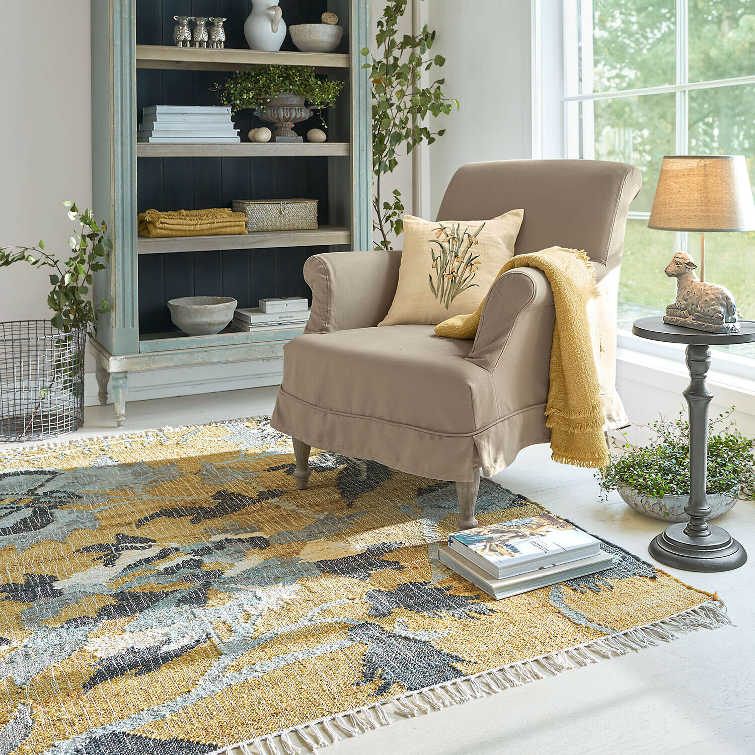 Beiger Polstersessel mit gelber Decke und Kissen auf buntem Teppich in hellem Wohnzimmer