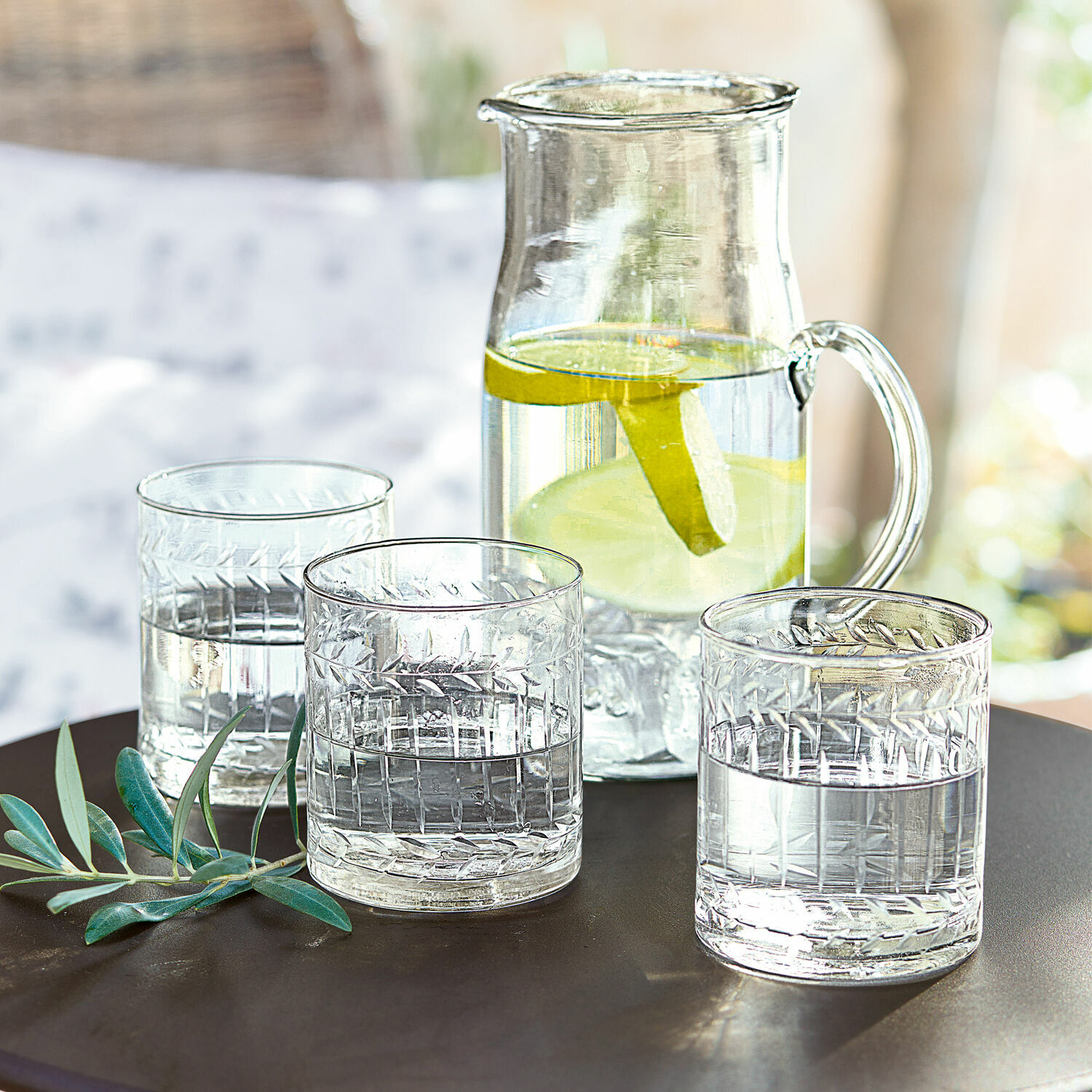 Glas 6er Set Padiès Glaskrug mit Zitronenscheiben und drei Wassergläser auf dunklem Tisch mit grünen Blättern als Dekoration