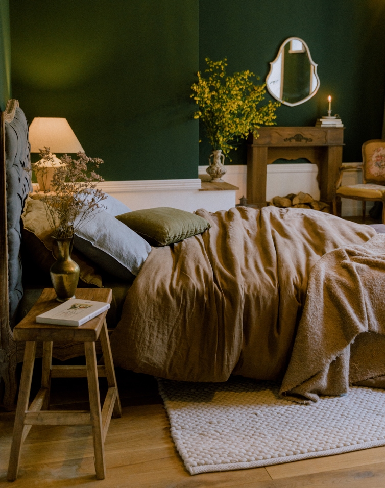 Gemütliches Schlafzimmer mit brauner Leinenbettwäsche und grünen Kissen vor dunkelgrüner Wand.