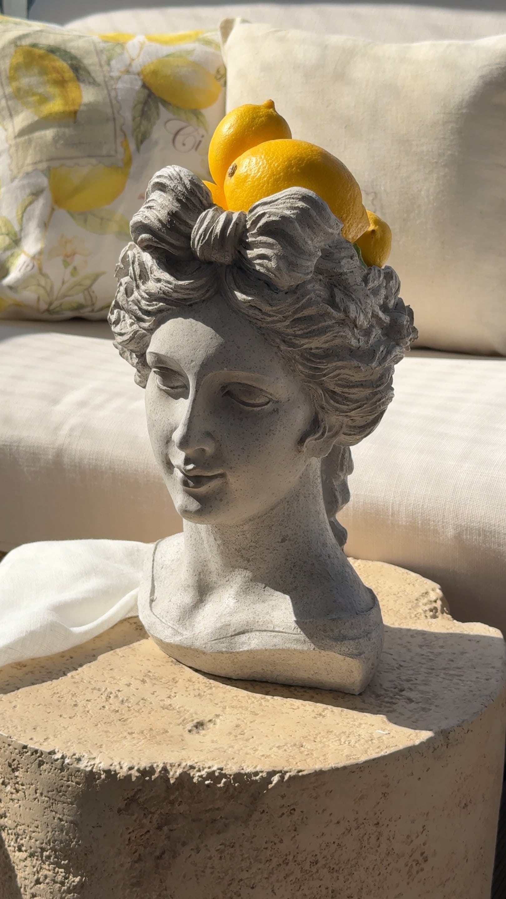 Graue Steinskulptur eines Frauenkopfs mit Zitronen auf dem Kopf vor hellen Kissen und Stoffen