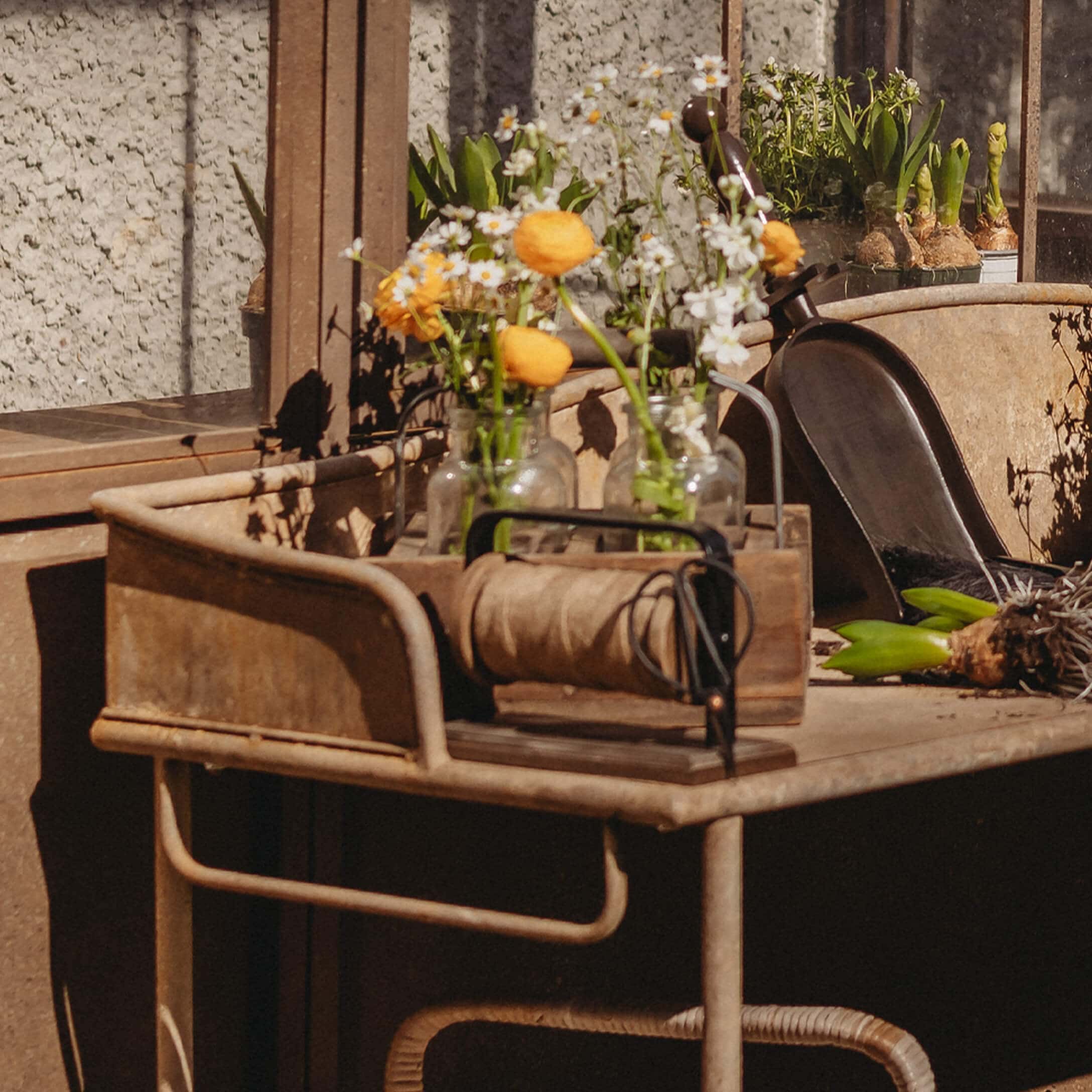 Metalltisch mit Glasvasen und gelben Blumen, Gartengeräte und Seilrolle auf sonnigem Balkon.