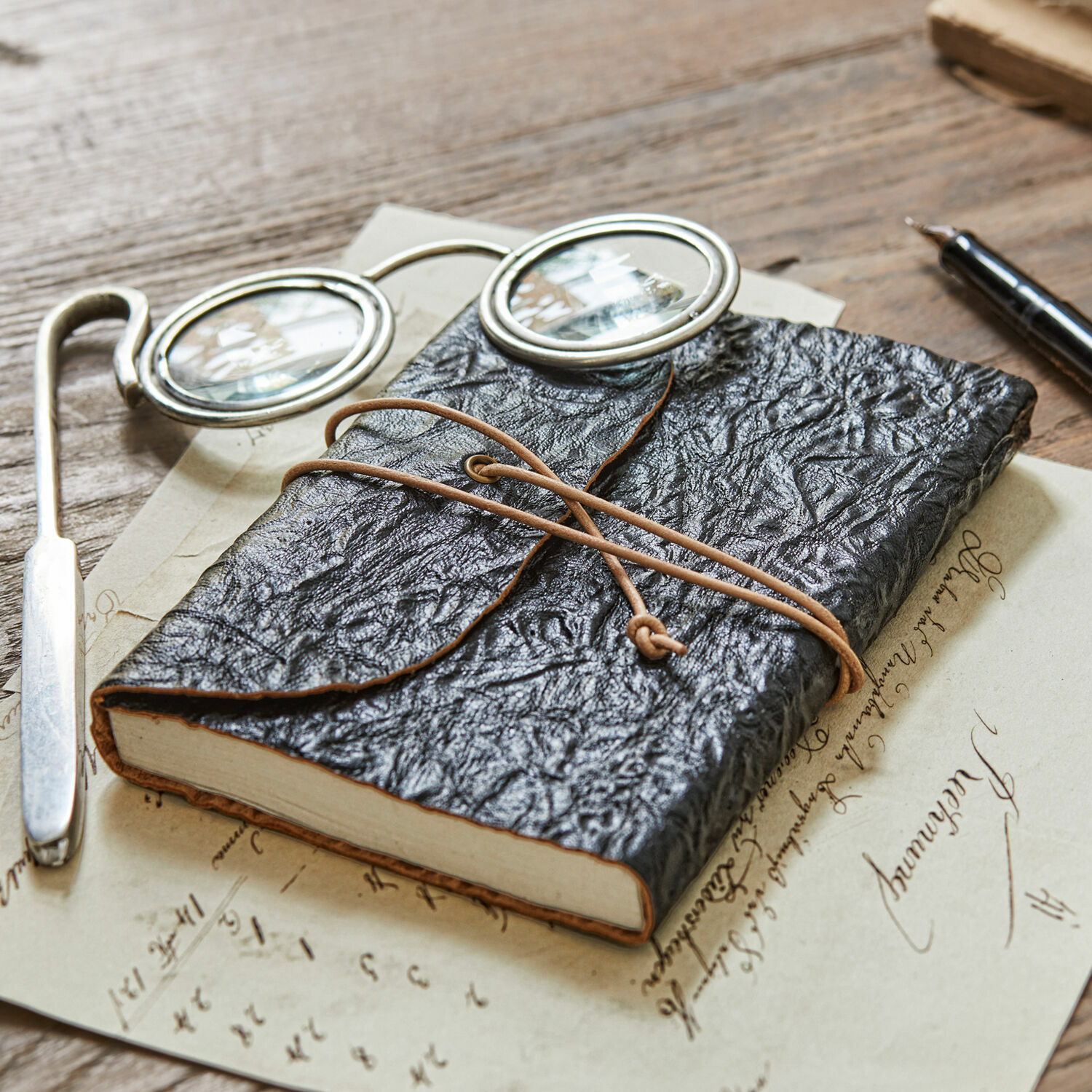 Notizbuch Cosmes Schwarzes Notizbuch aus strukturiertem Leder mit Lederriemen auf Holztisch mit Brille und Füllfederhalter