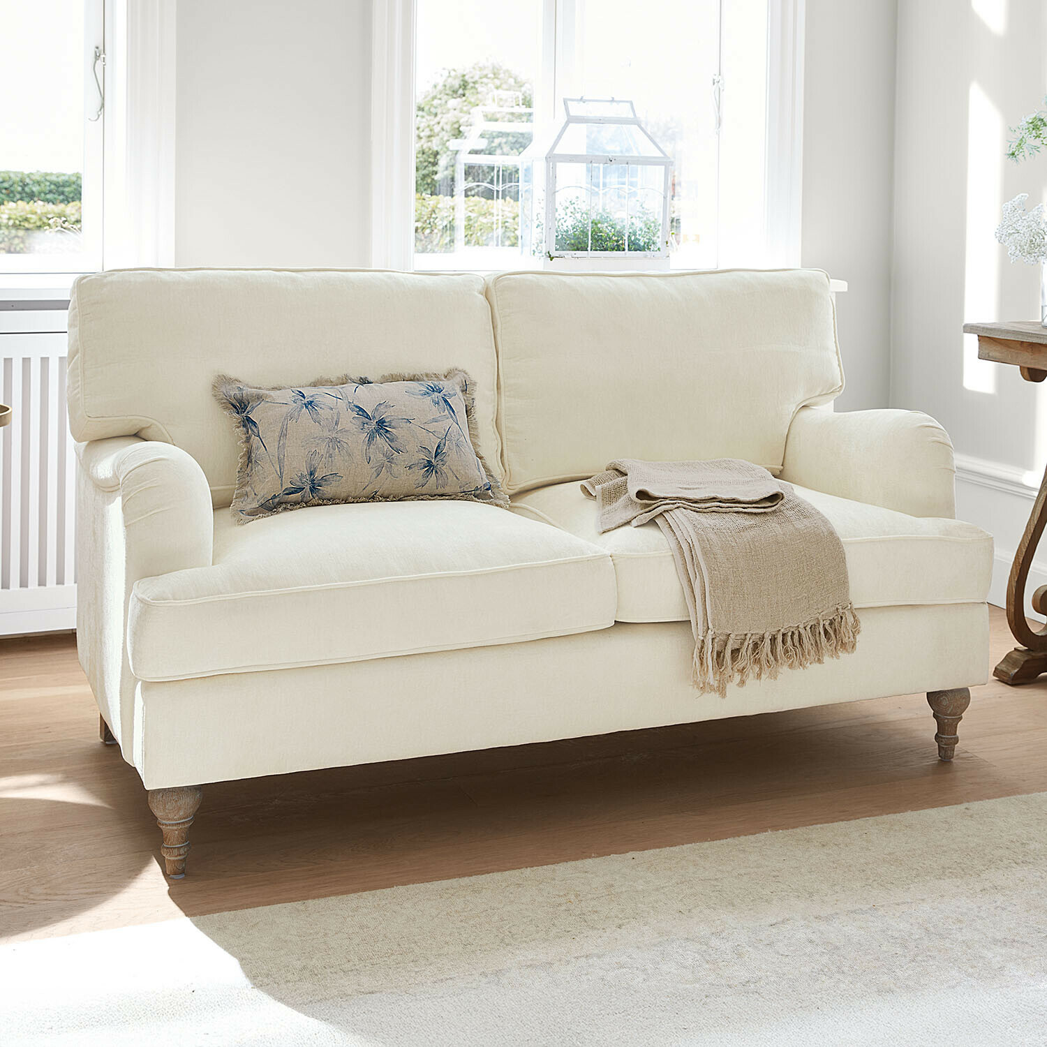 Sofa Elvika Beige bezogene Couch mit Blumen-Kissen und beigem Plaid in hellem Wohnzimmer