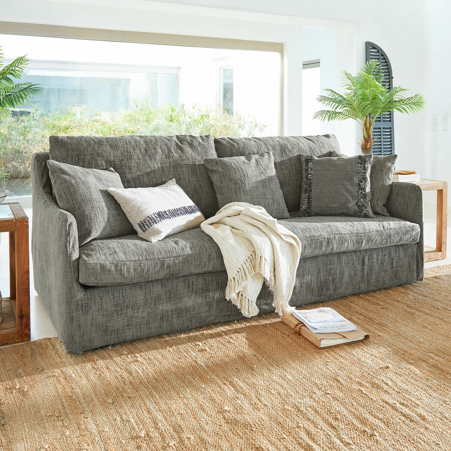 Sofa Shadowfield Graues Sofa aus Stoff mit Kissen und cremefarbener Decke auf Naturteppich im hellen Wohnzimmer