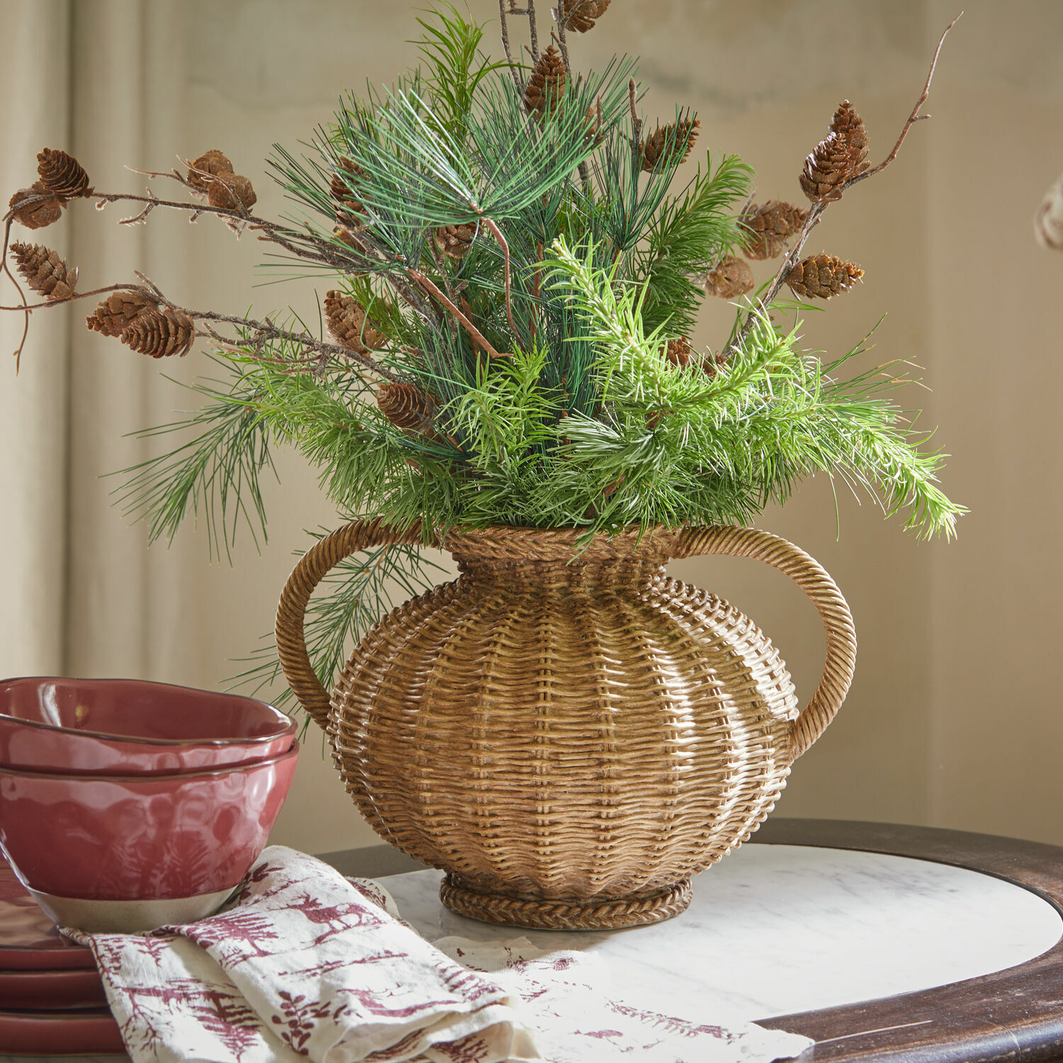 Vase Kaelvik Dekorativer Rattan-Vase mit Griffen, grünen Zweigen und Kiefernzapfen auf Tisch mit roter Keramik