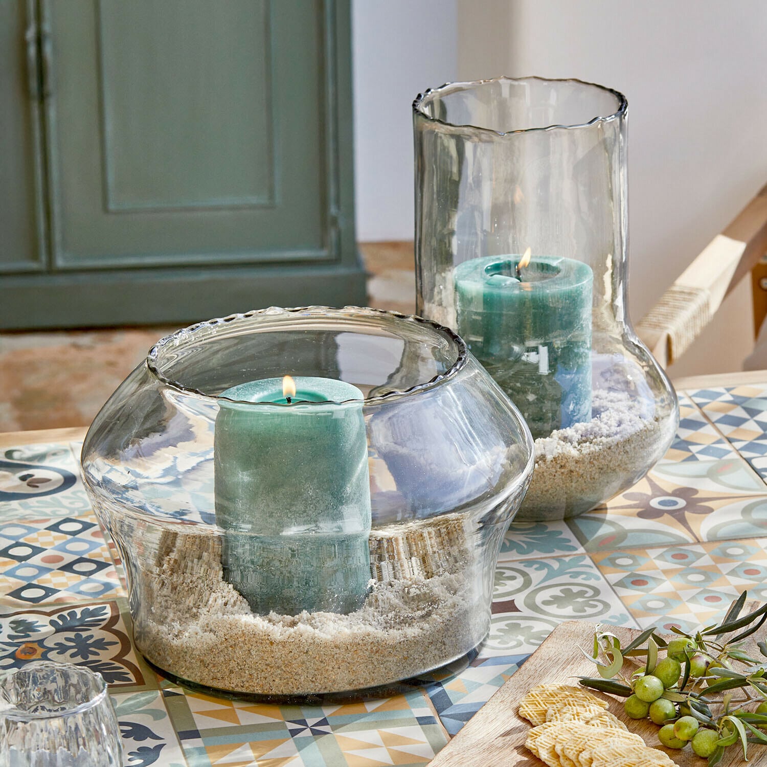 Windlicht 2er Set Lumiras Glaswindlichter mit grünen Kerzen und Sand auf einem bunten Tisch mit dekorativen Mustern