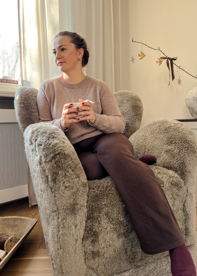 Eine Frau sitzt entspannt in einem grauen, kuscheligen Sessel aus Fellimitat und hält eine Tasse in der Hand.