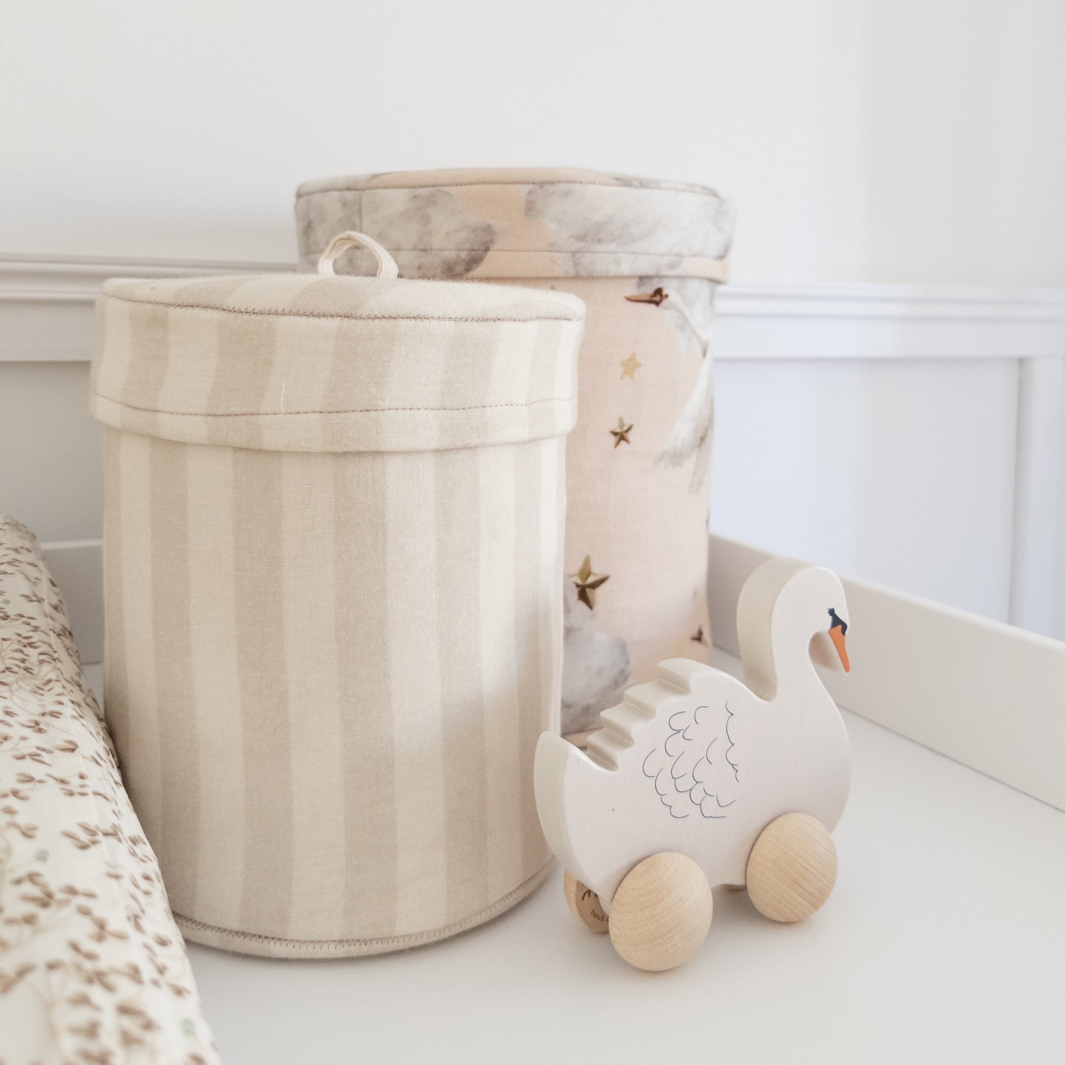 Zwei Stoffbehälter in Beige mit Streifen und Mustern sowie ein hölzernes Schwan-Spielzeug auf Rädern fürs Kinderzimmer