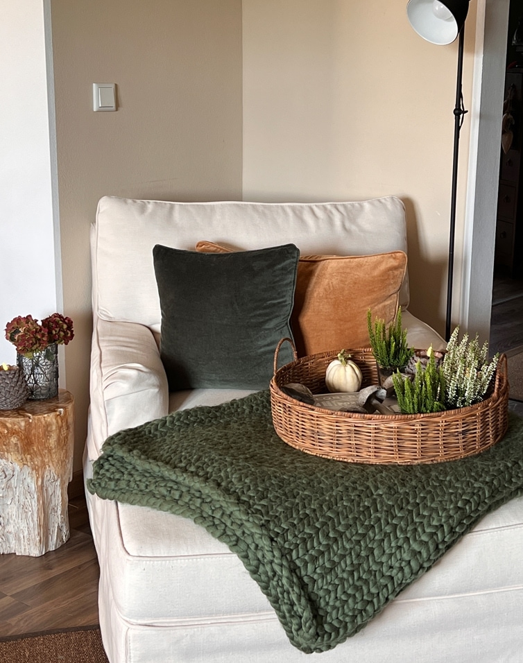 Beiger Sessel mit grünem und orangefarbenem Kissen, Korb mit Dekorationen auf grünem Strickplaid.