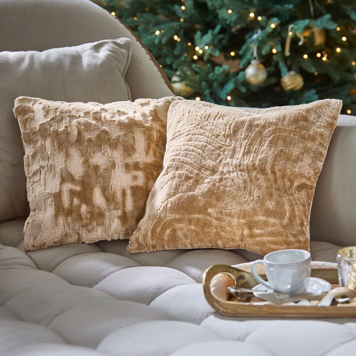 Zwei beige dekorative Kissen aus weichem Stoff auf hellem Sofa vor geschmücktem Weihnachtsbaum