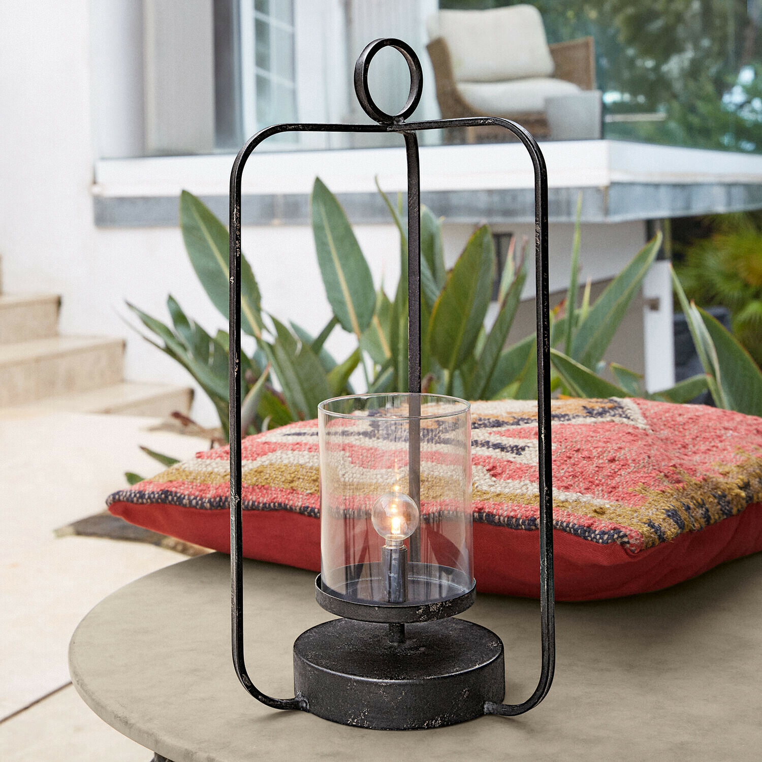 LED-Laterne Kivalex Schwarze Metalllaterne mit klarem Glas auf Gartentisch neben buntem Kissen