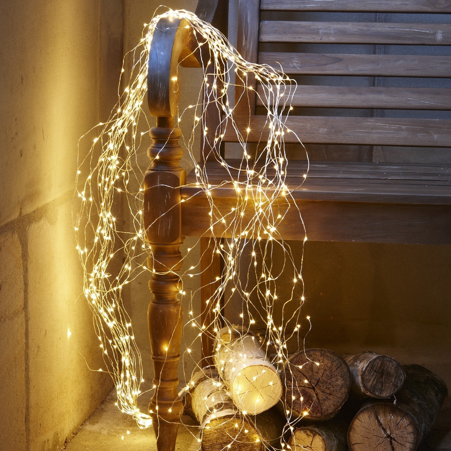 Lichterkette Voltage Feine warmweisse LED-Lichterkette auf dünnen Drähten um eine Holzbank und Holzscheite gewickelt
