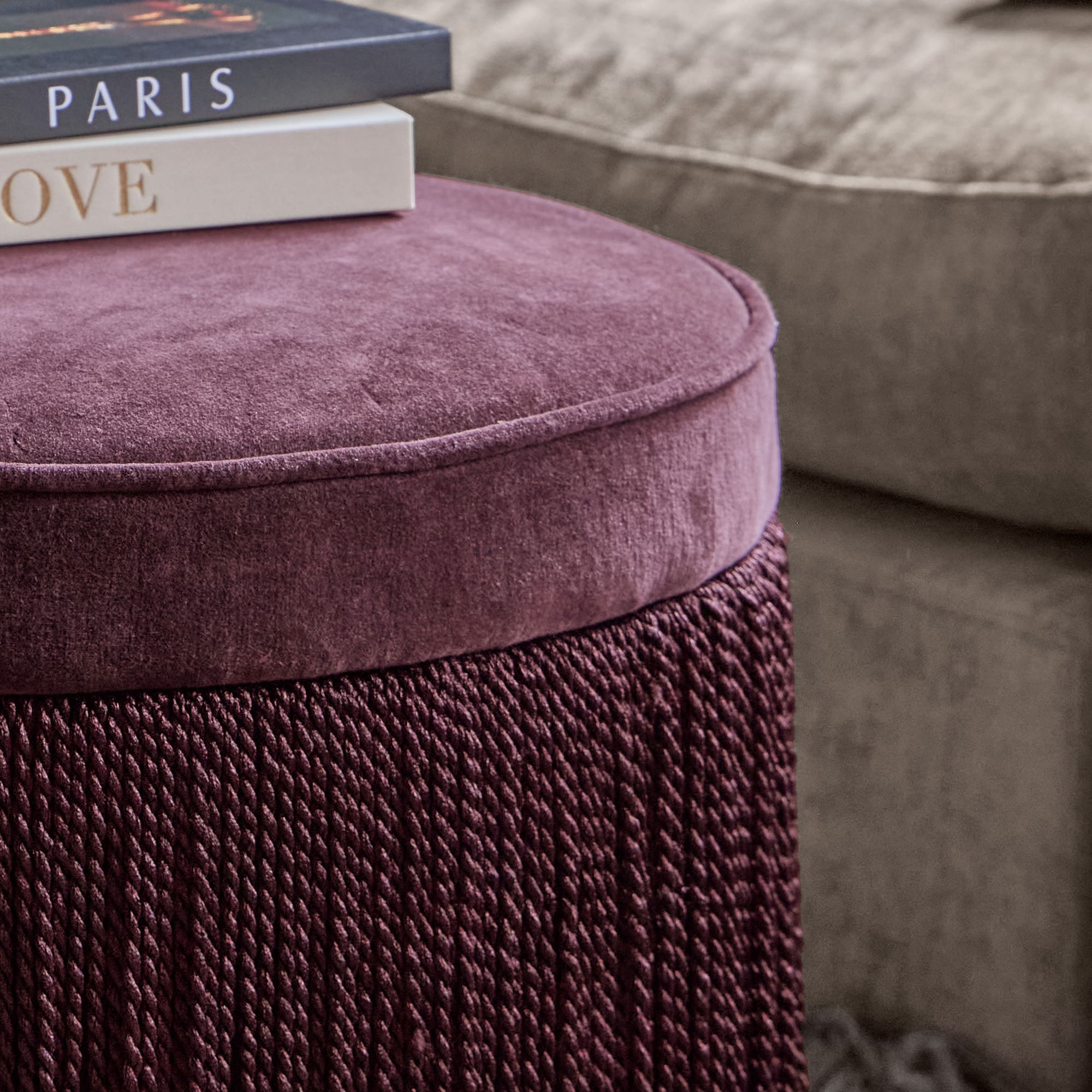 Violetter Samt-Pouf mit geflochtenem Stoff vor grauem Sofa und Büchern im Hintergrund