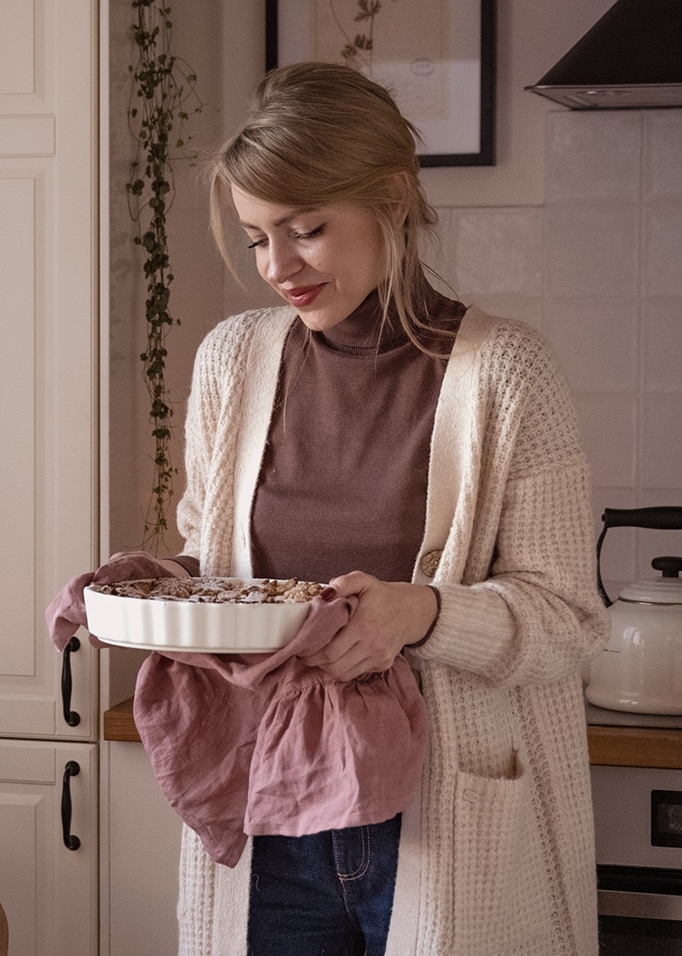 Frau im beigen Strickcardigan hält weisse Keramik-Backform mit rosa Tuch in der Küche.