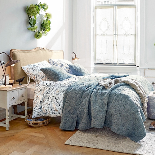 Gemütliches Schlafzimmer mit blauem Steppbett und gemustertem Bettwäscheset an beigem Kopfteil