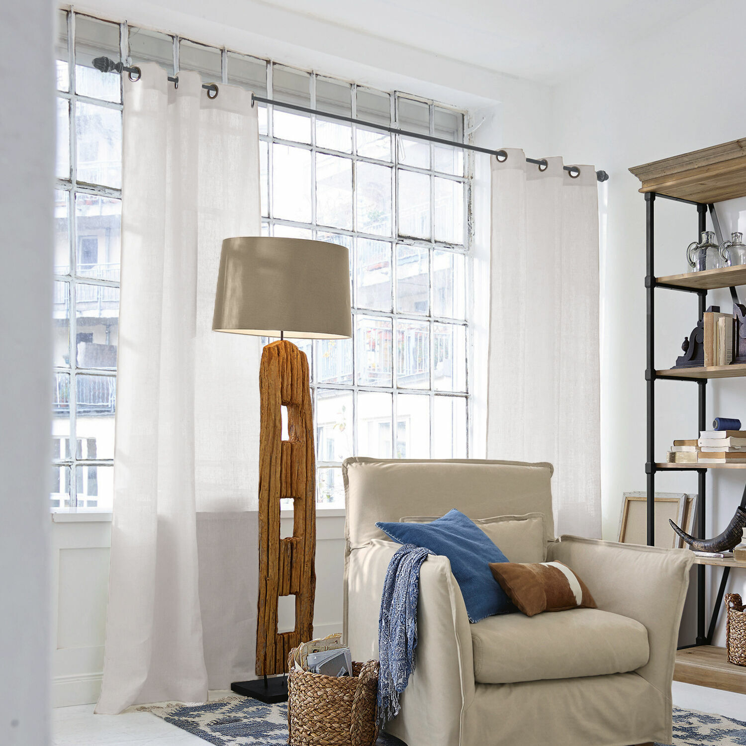 Gardine Salinas Stehlampe mit Holzfuss und beigem Lampenschirm in hellem Wohnzimmer mit Sessel und Regal
