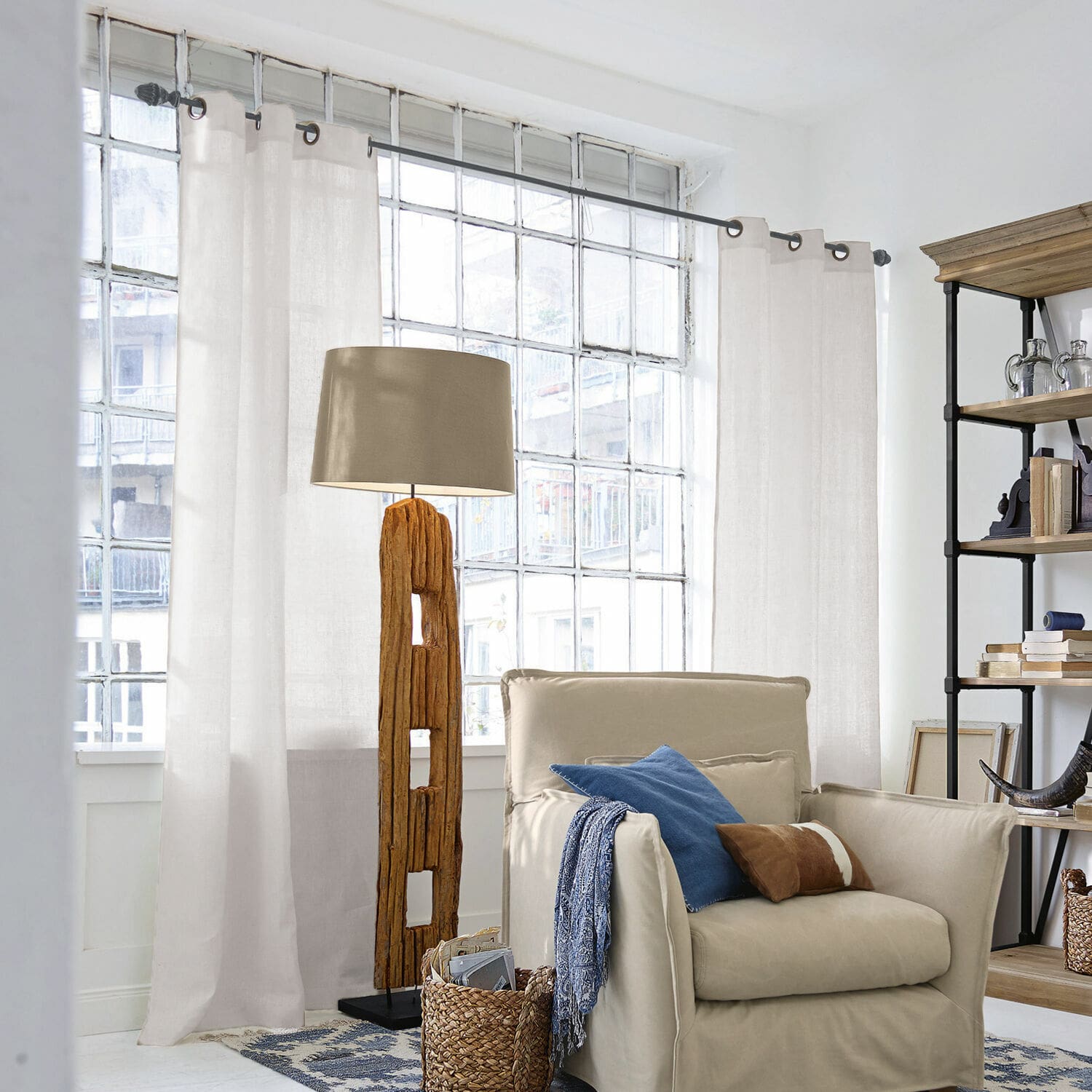 Gardine Salinas Stehlampe mit Holzfuss und beigem Lampenschirm in hellem Wohnzimmer mit Sessel und Regal