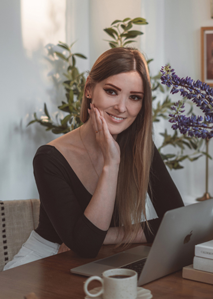 Frau sitzt an Holztisch mit Laptop, Kaffeetasse und Blumen im Hintergrund.