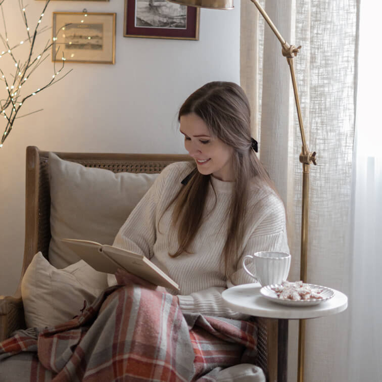 Frau liest Buch auf Korbsessel mit cremefarbenem Pullover und warmem kariertem Plaid