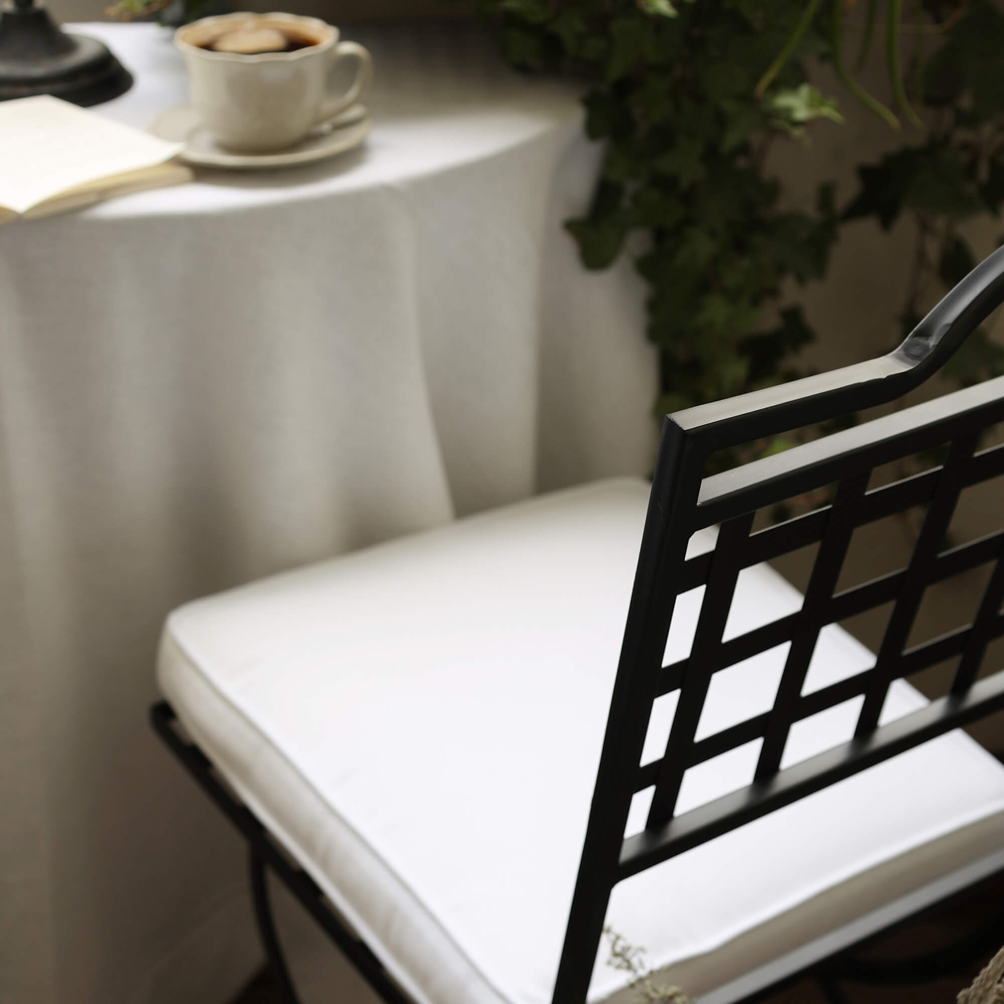 Schwarzer Metallstuhl mit weissem Sitzkissen vor Tisch mit Kaffeetasse und grünen Pflanzen auf dem Balkon