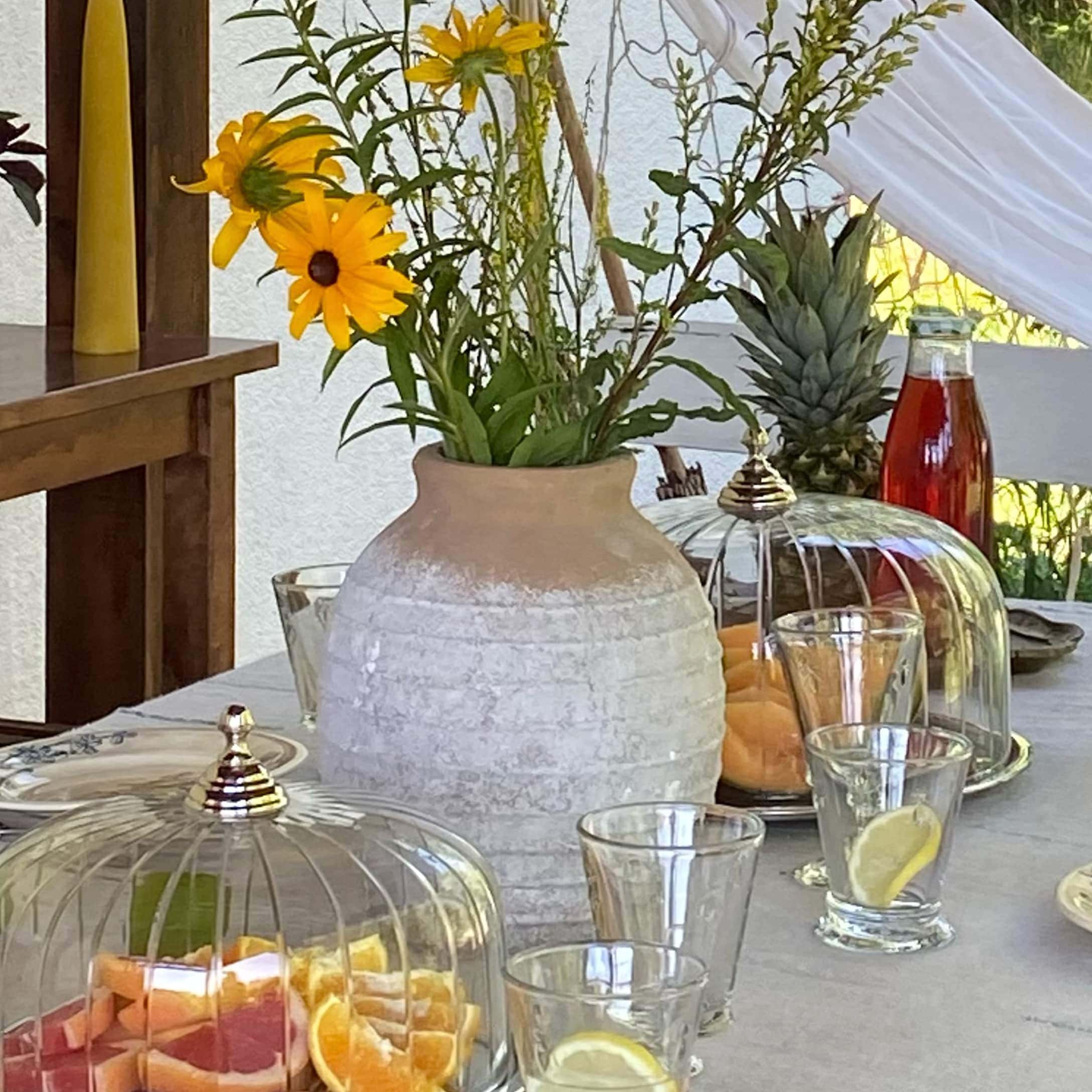 Keramikvase in Beige-Weiss mit gelben Blumen auf Tisch mit Früchten und Gläsern