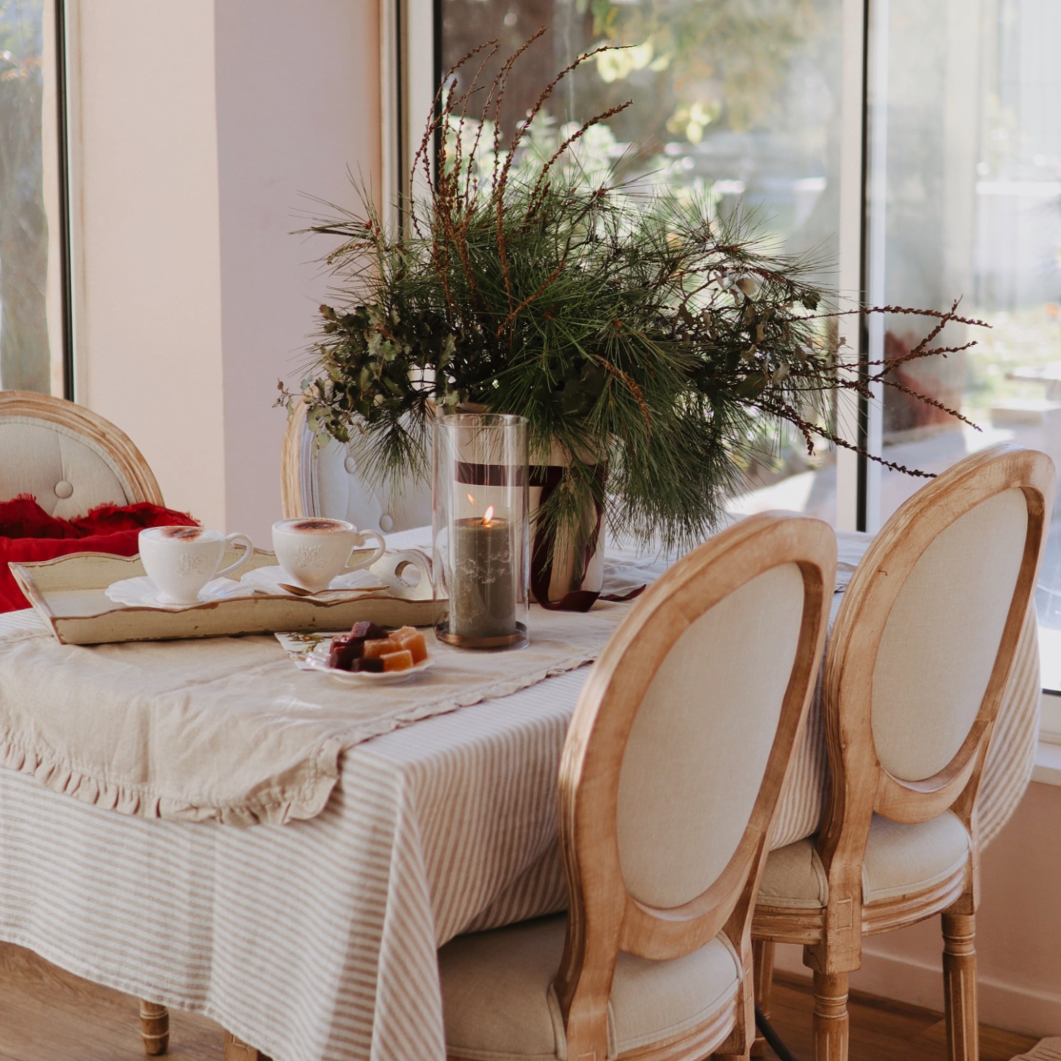 Gemütlicher Tisch mit beigem Tischläufer, Holzstühlen und weihnachtlicher Dekoration aus grünen Zweigen.