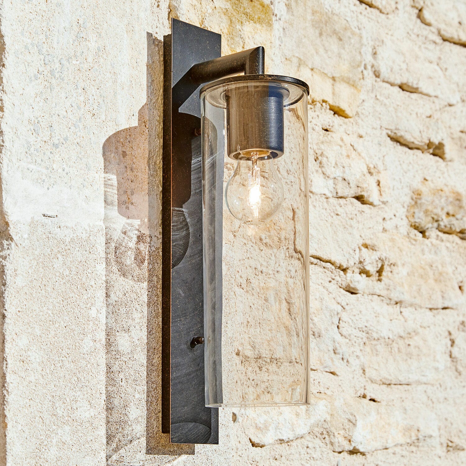 Aussenwandlampe Clisson Schwarze Metall-Wandleuchte mit klarem Glaszylinder an heller Steinwand für den Aussenbereich