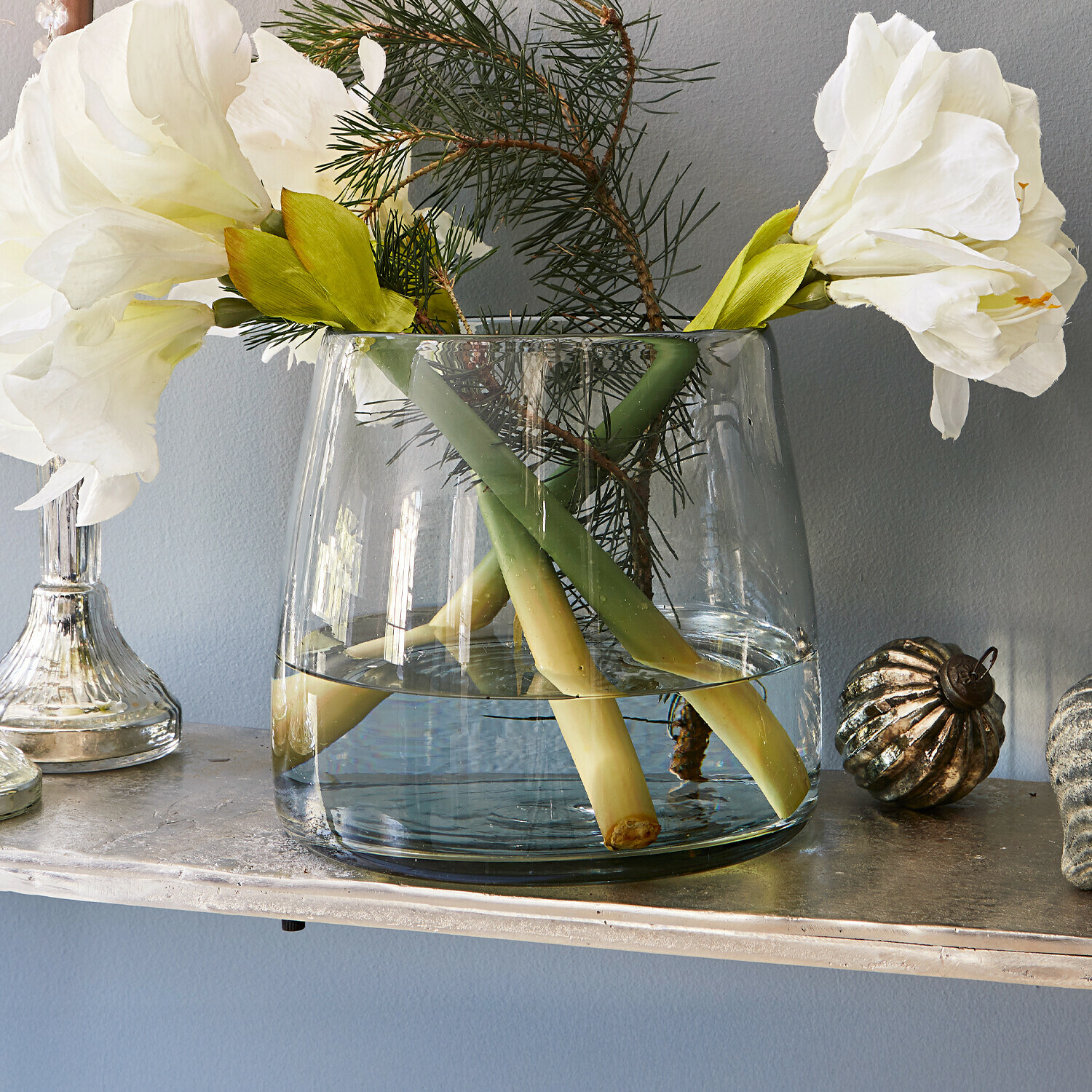 Vase Inga Transparente Glasvase mit weissen Blumen und grünen Zweigen auf einem dekorativen Regal.