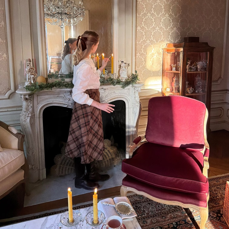 Frau zündet Kerzen an einem Marmorkamin in elegantem Wohnzimmer mit bordeauxrotem Sessel an