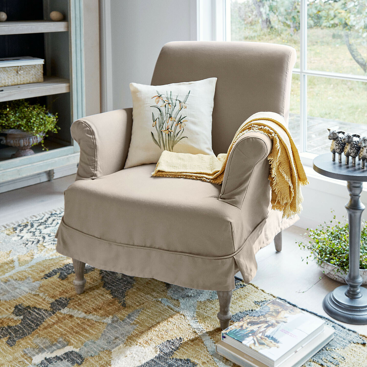 Sessel Everleighs Beiger Stoffsessel mit Blumen-Kissen und gelbem Plaid in hellem Wohnzimmer mit Teppich