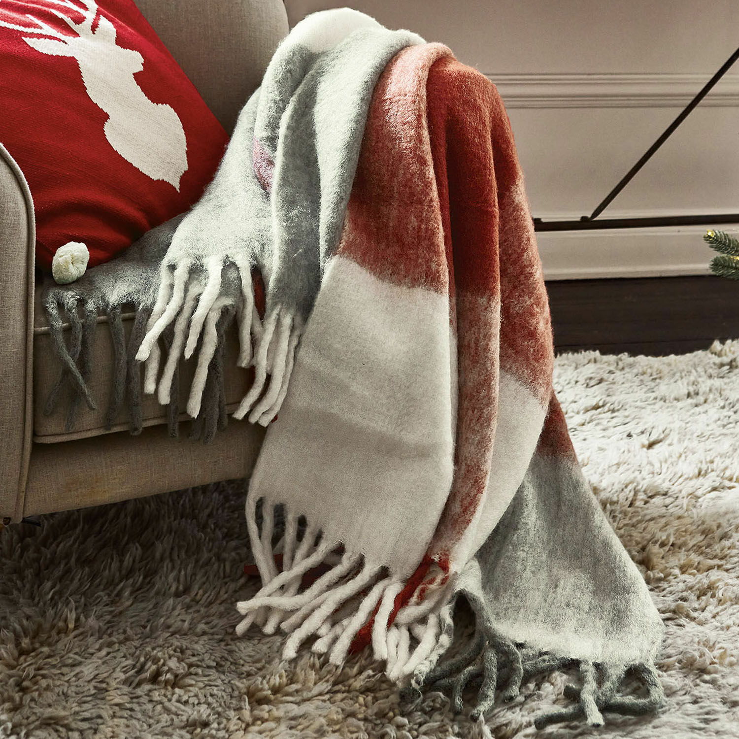 Plaid McGreg Weiche Kuscheldecke mit Fransen in Grau, Weiss und Rot auf beigem Sofa, gemütliche Wohnraumdekoration