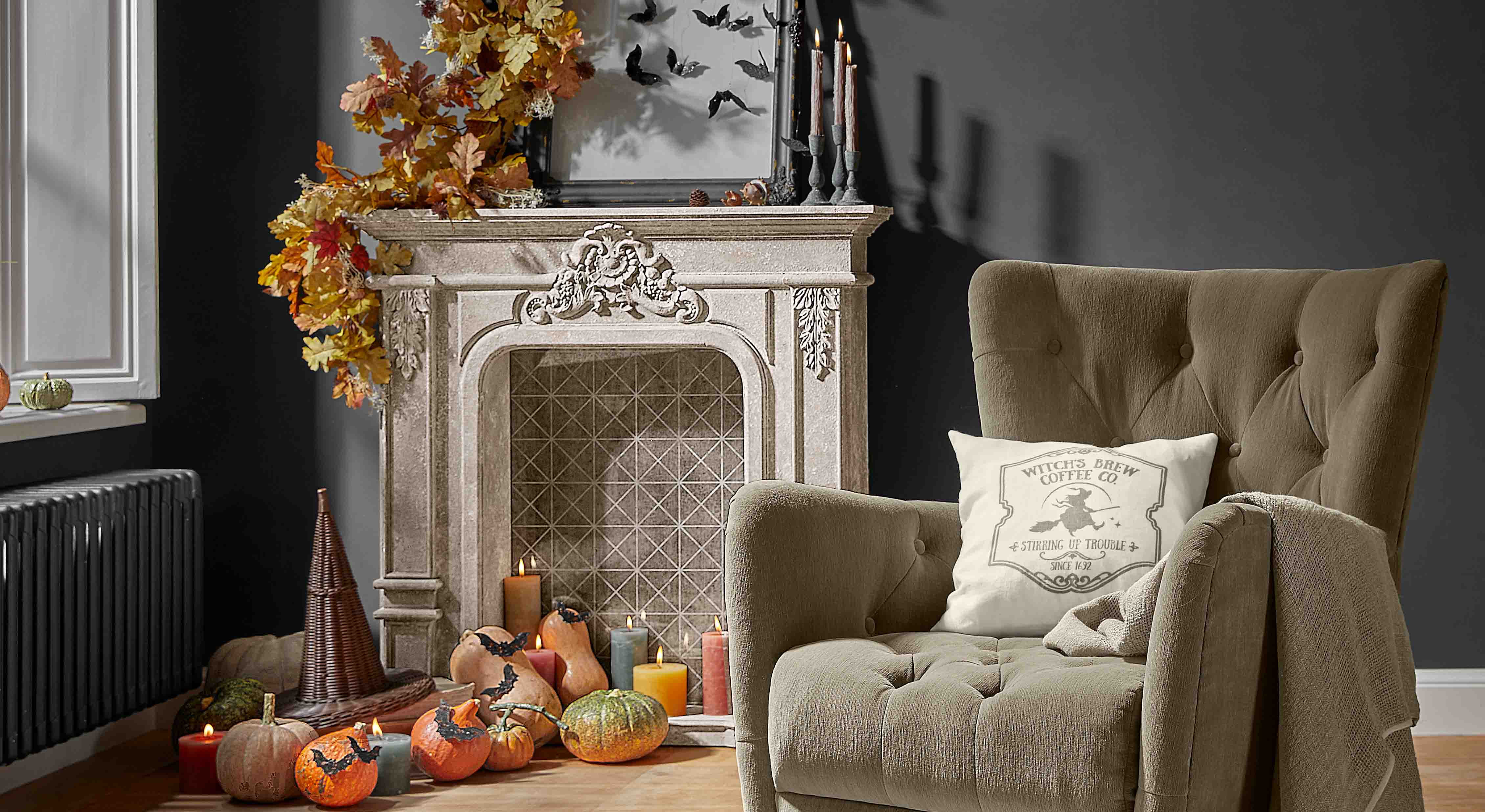 Dekorativer Kamin mit Herbstlaub, Kürbissen und Kerzen in gemütlichem Wohnzimmer mit beigem Sessel