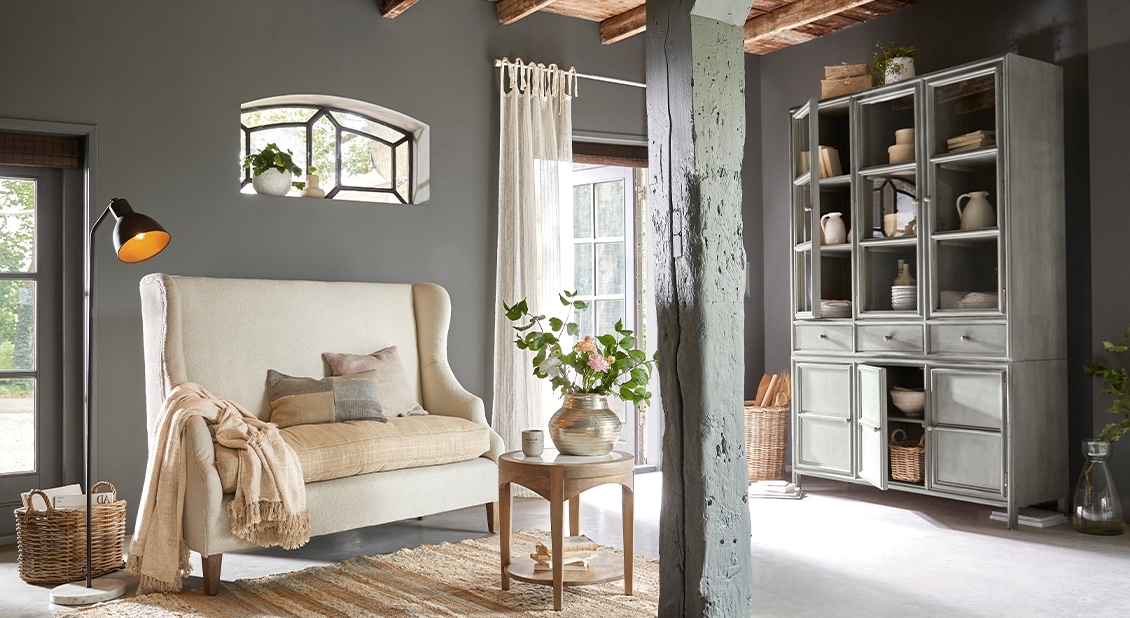 Gemütliches Wohnzimmer mit grauem Holz-Vitrinenschrank und cremefarbenem Sofa auf Holzbeinen, natürliche Dekoration