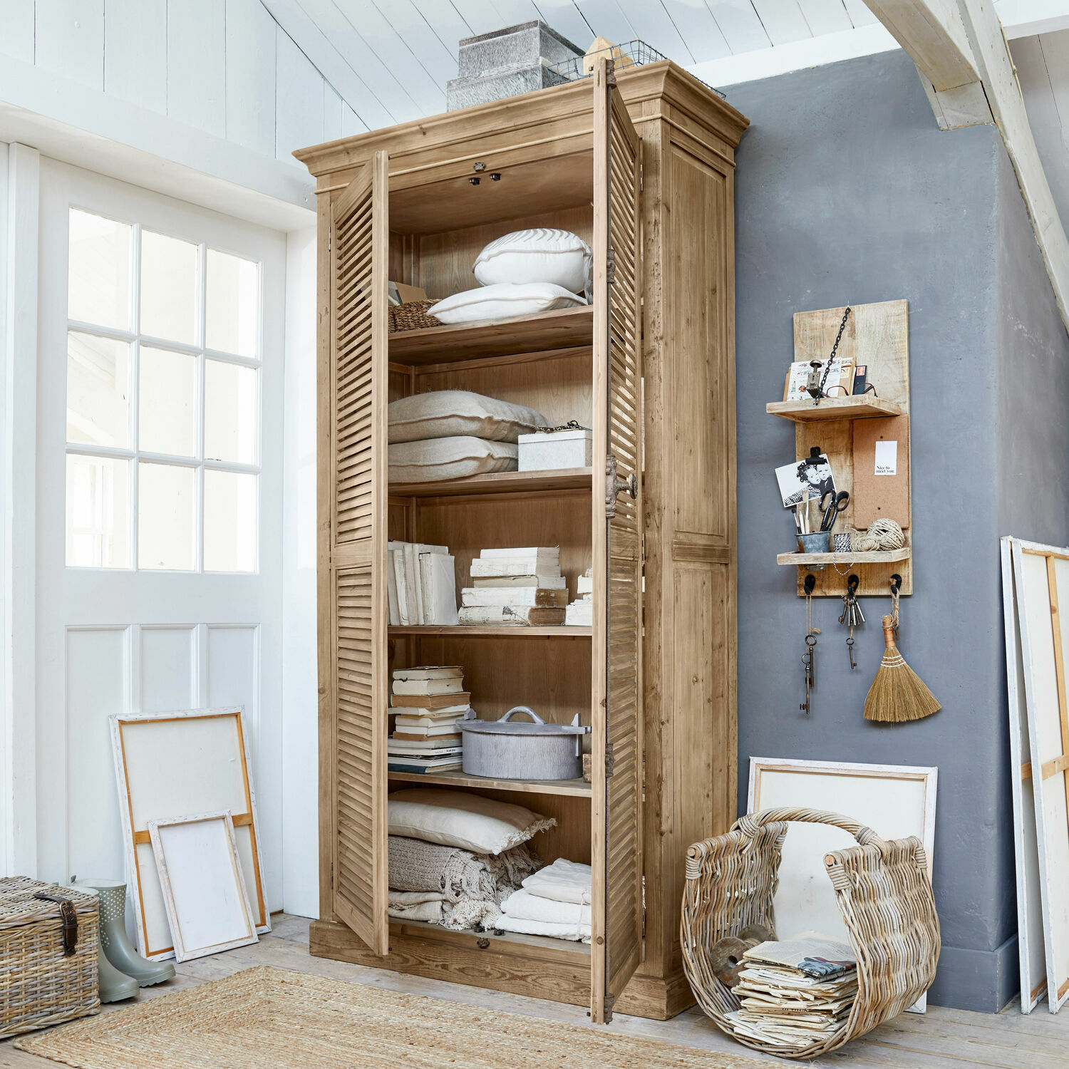 Schrank Farnham Holzschrank mit Lamellentüren in Naturholzfarbe, gefüllt mit Kissen und Büchern in hellem Raum.