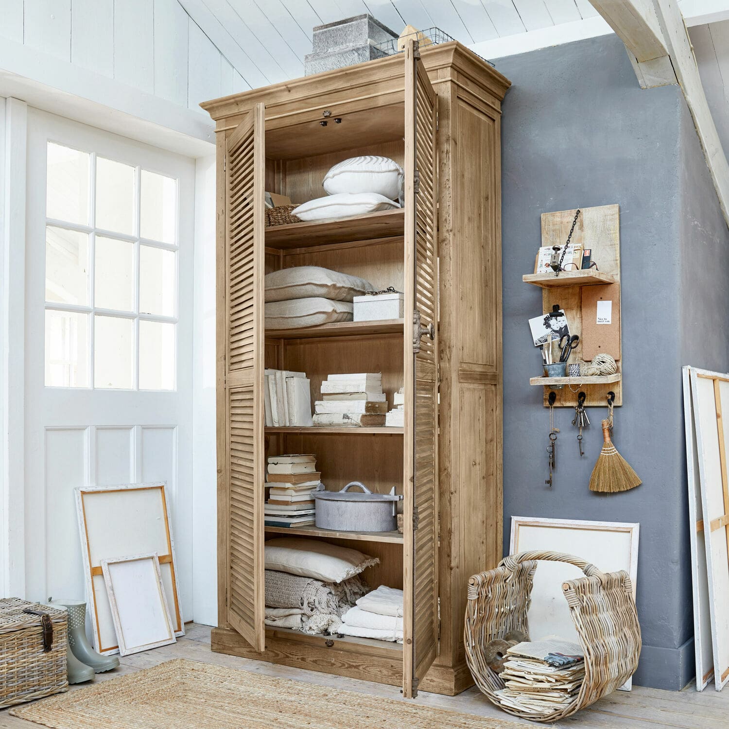 Schrank Farnham Holzschrank mit Lamellentüren in Naturholzfarbe, gefüllt mit Kissen und Büchern in hellem Raum.