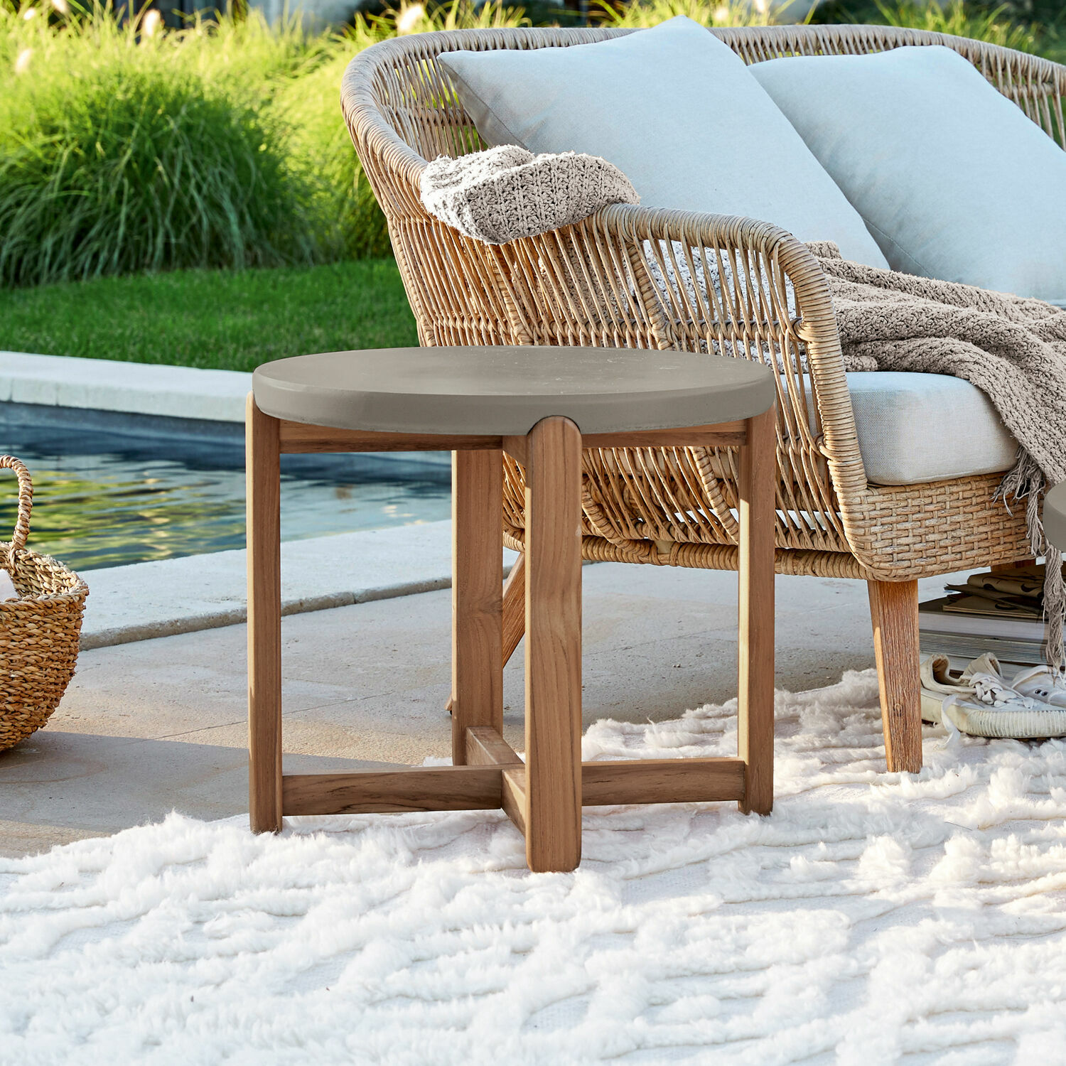 Beistelltisch Robinson Runder Beistelltisch mit Holzgestell und grauer Tischplatte auf der Terrasse beim Pool mit Teppich und Korbsessel