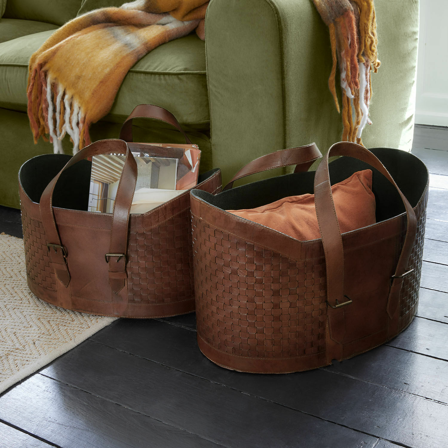 Korb 2er Set Tavari Braune Ledertaschen mit Flechtmuster zur Aufbewahrung neben grünem Sofa auf dunklem Boden