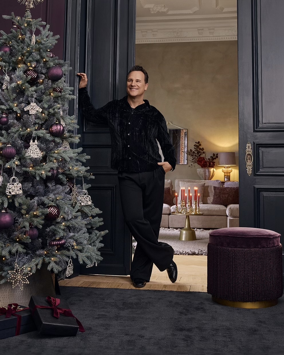 Mann in schwarzer Kleidung lehnt an Türrahmen neben Weihnachtsbaum mit violetten Kugeln und rotem Kerzenständer im Wohnzimmer