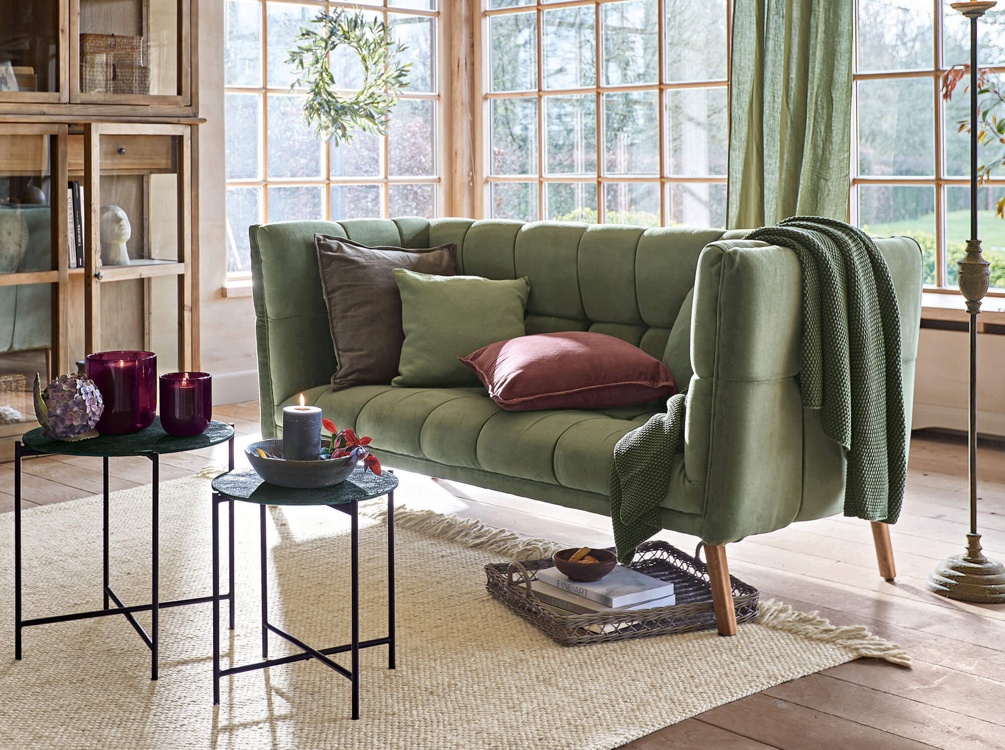 Grünes Samt-Vintage-Sofa mit Kissen und Decke in hellem Wohnzimmer mit grossen Fenstern
