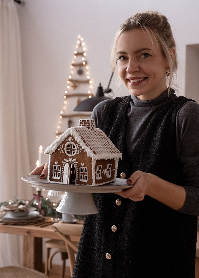 Eine Frau hält einen Teller mit einem Lebkuchenhaus in einem gemütlich weihnachtlich dekorierten Raum.