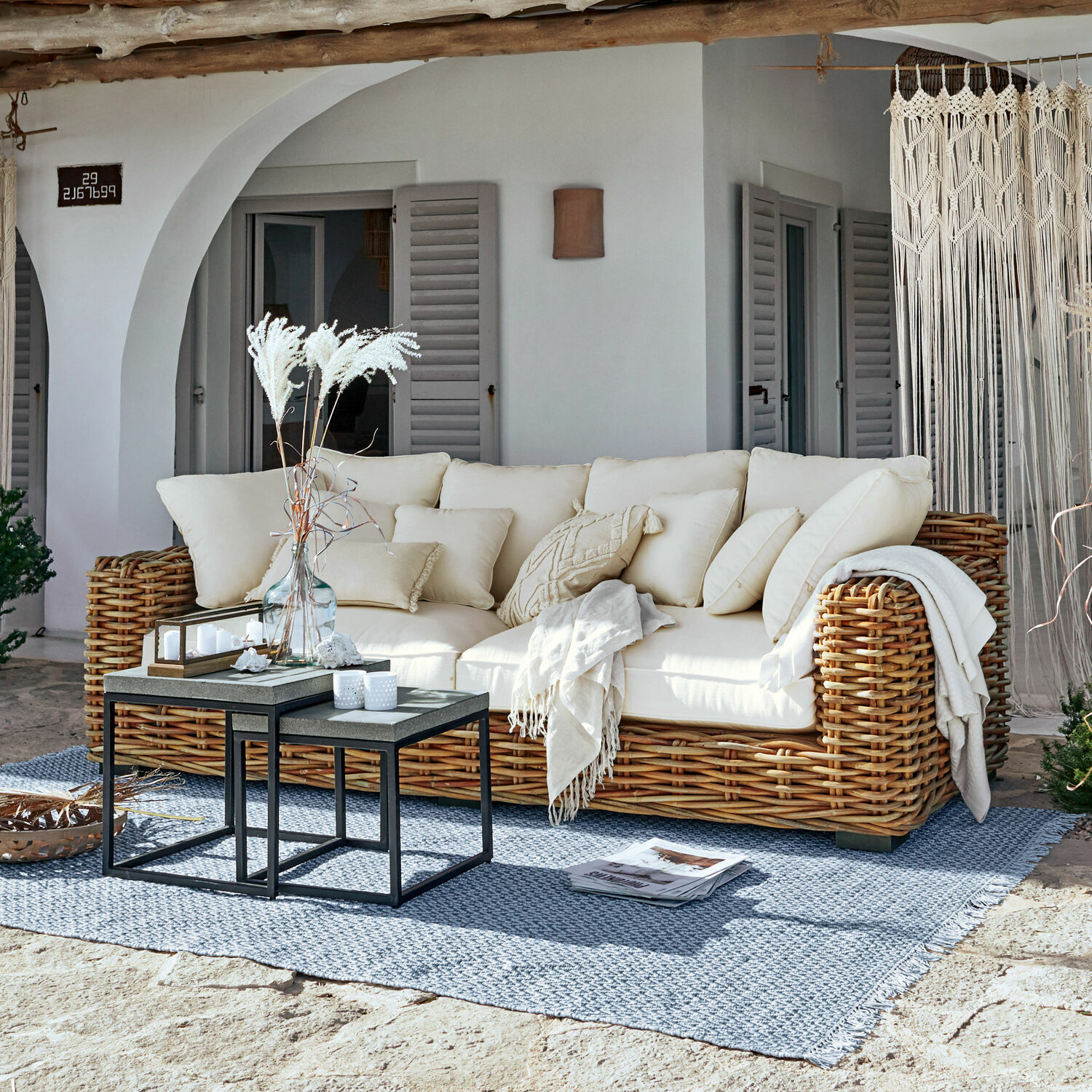 Sofa Rimini Rattansofa mit cremefarbenen Kissen und Decke auf blauem Teppich auf der sonnigen Terrasse