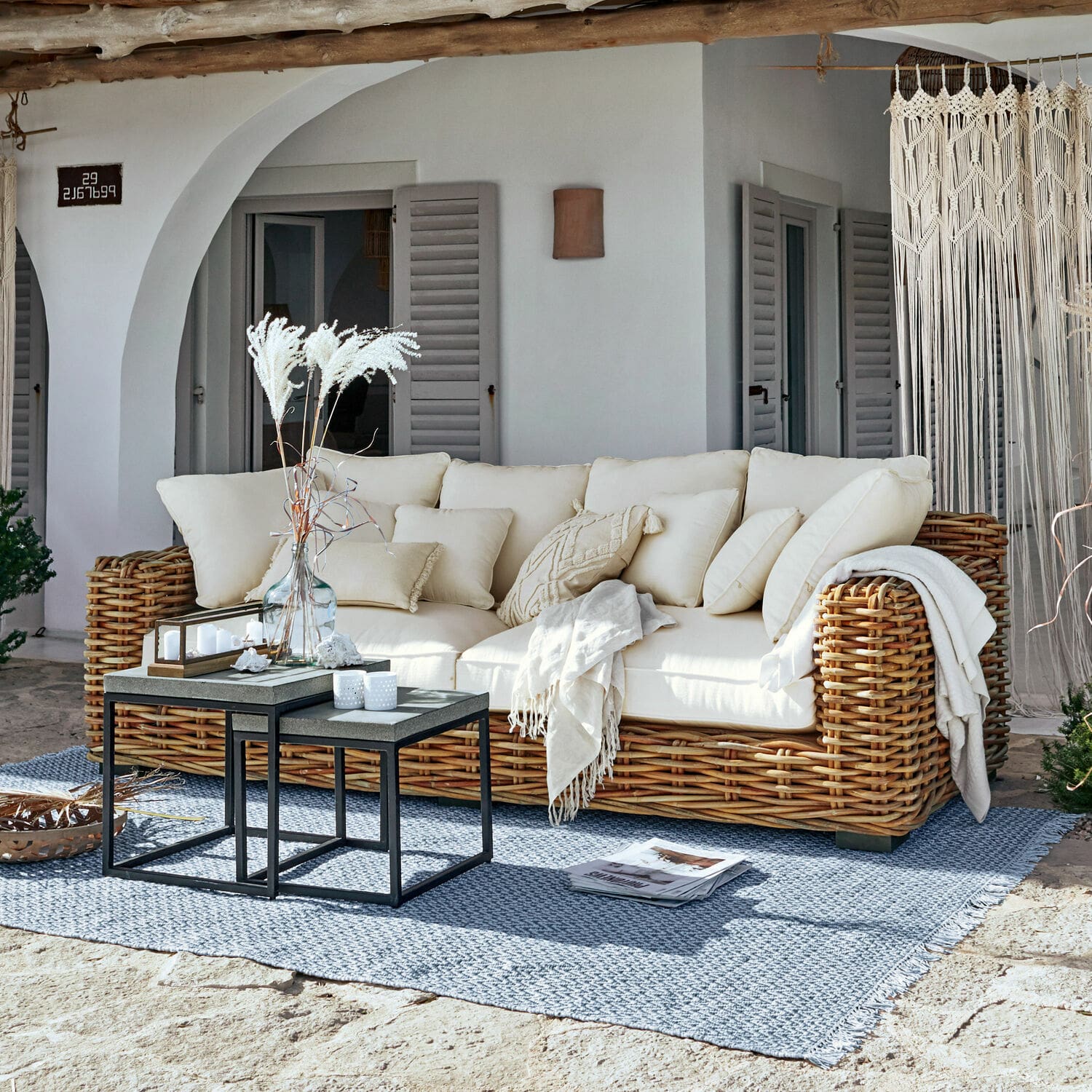 Sofa Rimini Rattansofa mit cremefarbenen Kissen und Decke auf blauem Teppich auf der sonnigen Terrasse