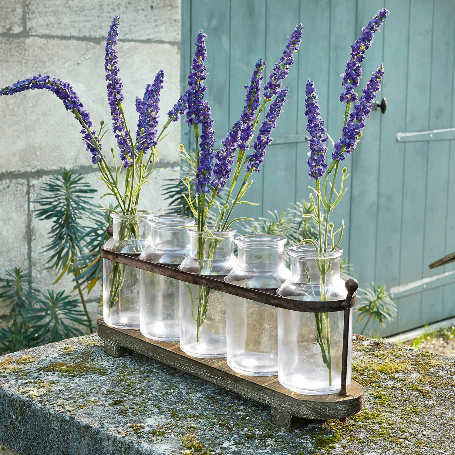 Vase 5er Set Ranjo Set aus fünf transparenten Glasvasen mit Metallhalterung auf Holzsockel, dekoriert mit violetten Blumen.
