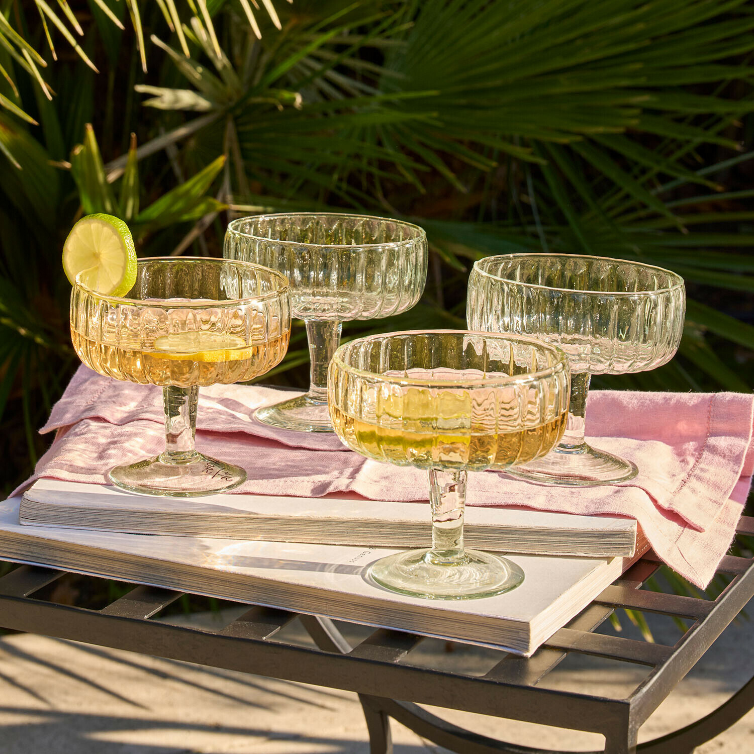 glas 4er Set Solerón Set mit vier transparenten Cocktailgläsern aus geriffeltem Glas auf Metalltisch mit Buch und rosa Serviette