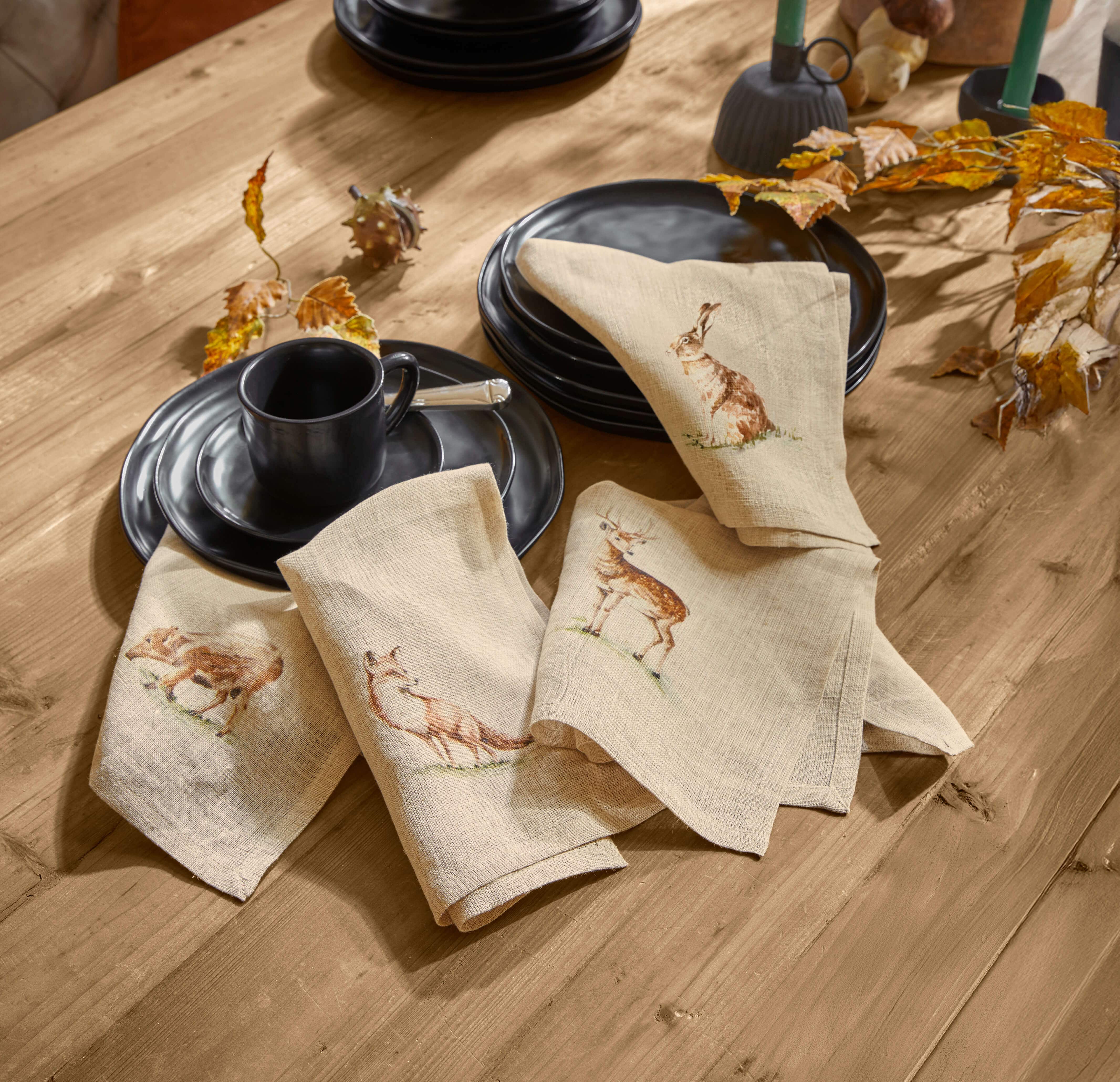 Beige Leinenservietten mit Waldtier-Motiven auf Holztisch neben schwarzem Geschirr