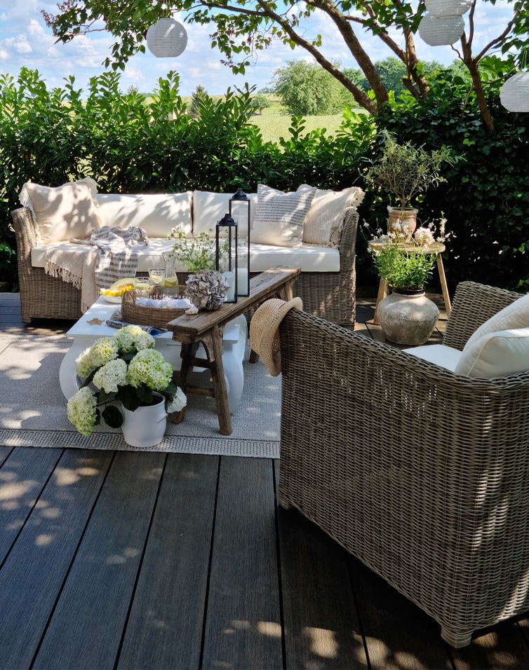 Gemütliche Gartenmöbel aus Rattan mit beigen Kissen auf der Terrasse, dekoriert mit Blumen und Laternen